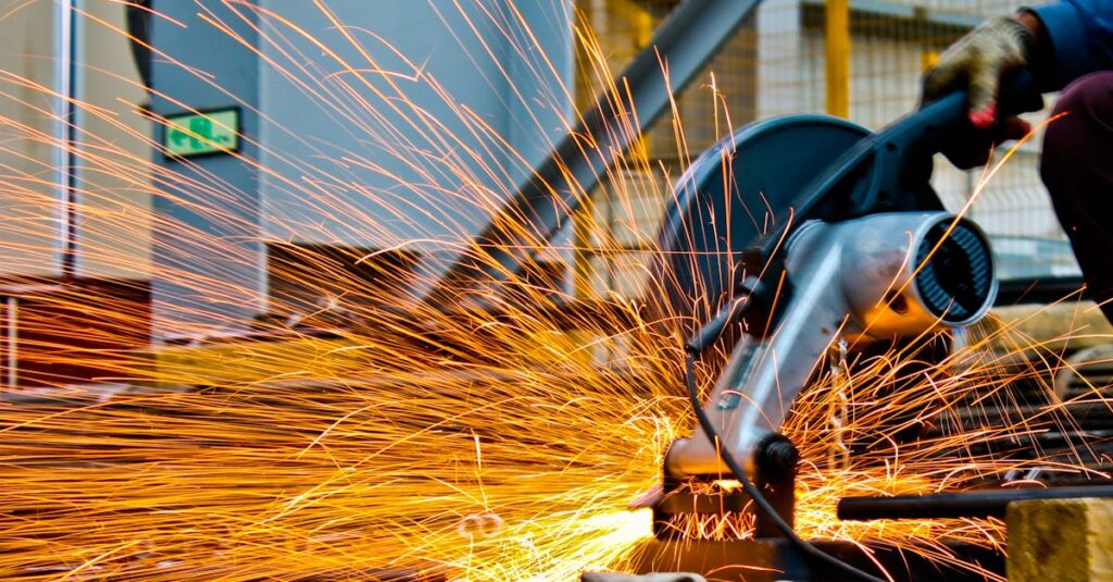 A worker operates a grinder cutting metal, creating a vibrant display of sparks in an industrial setting.